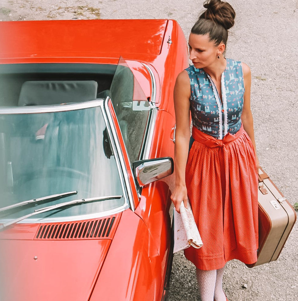 Eine Frau steht neben einem roten Cabrio und trägt ein Dirndl.